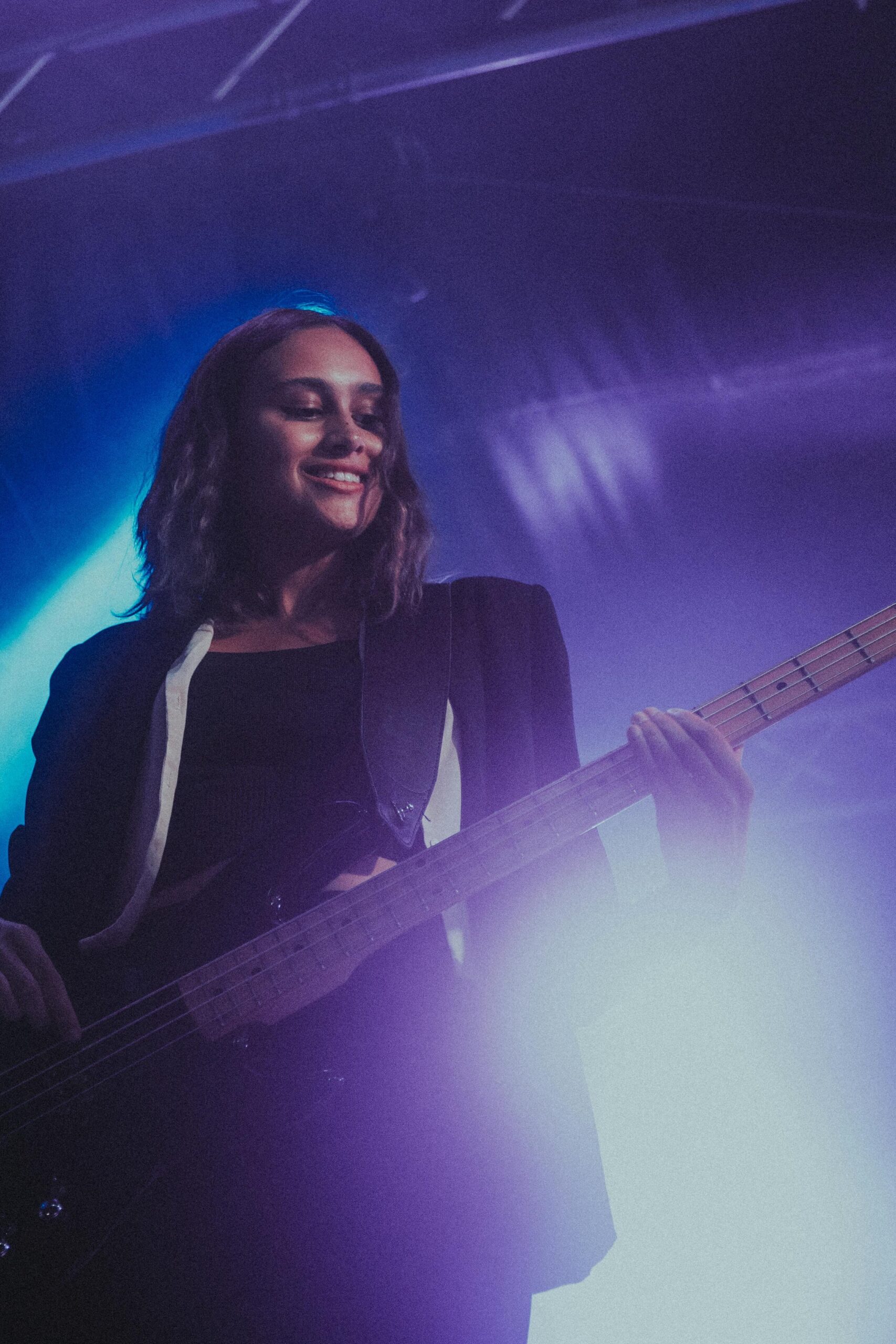 Woman playing bass guitar on stage with moody, vibrant lighting during a live performance.