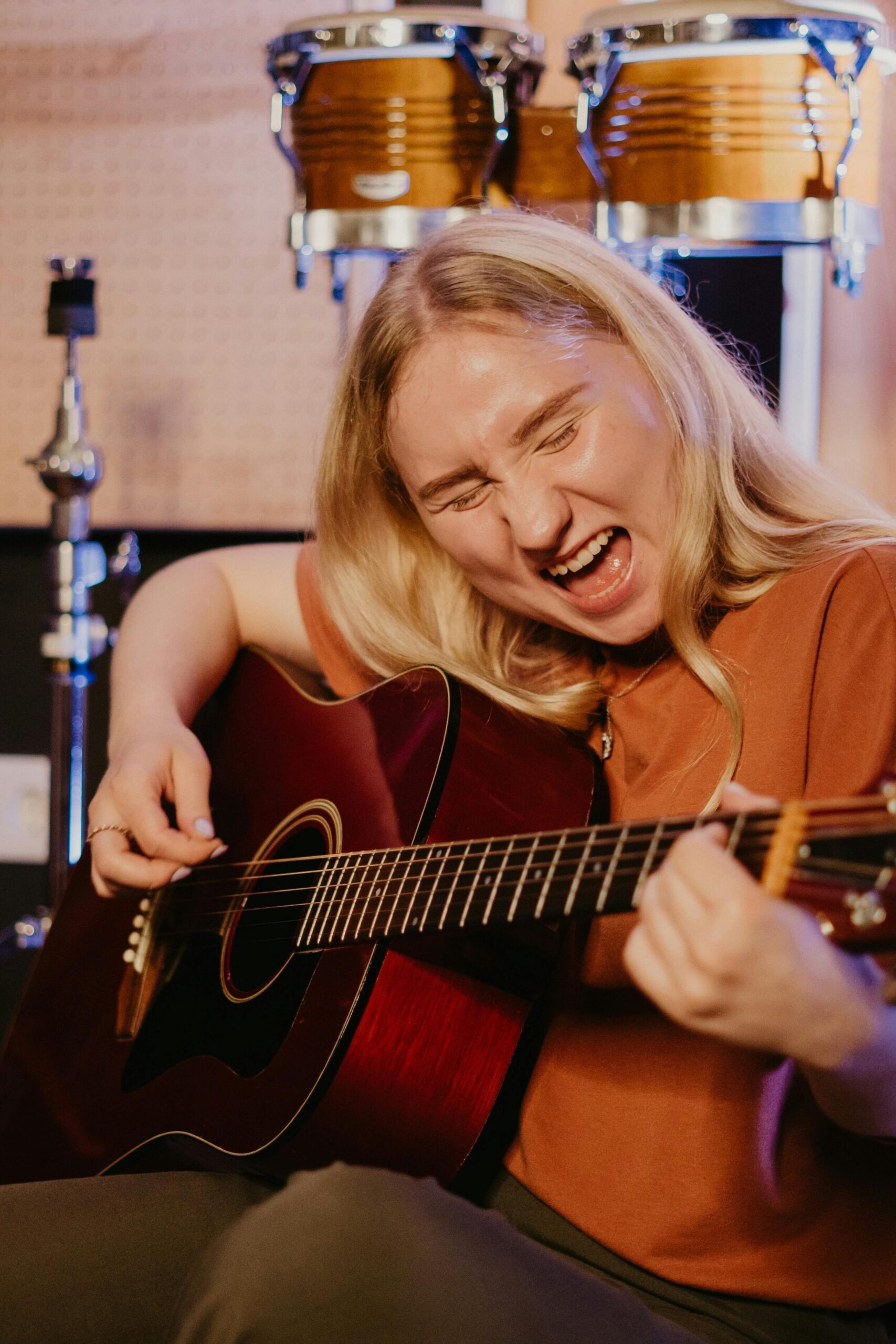 Blonde woman intensely playing acoustic guitar with drums in background, showcasing musical passion.