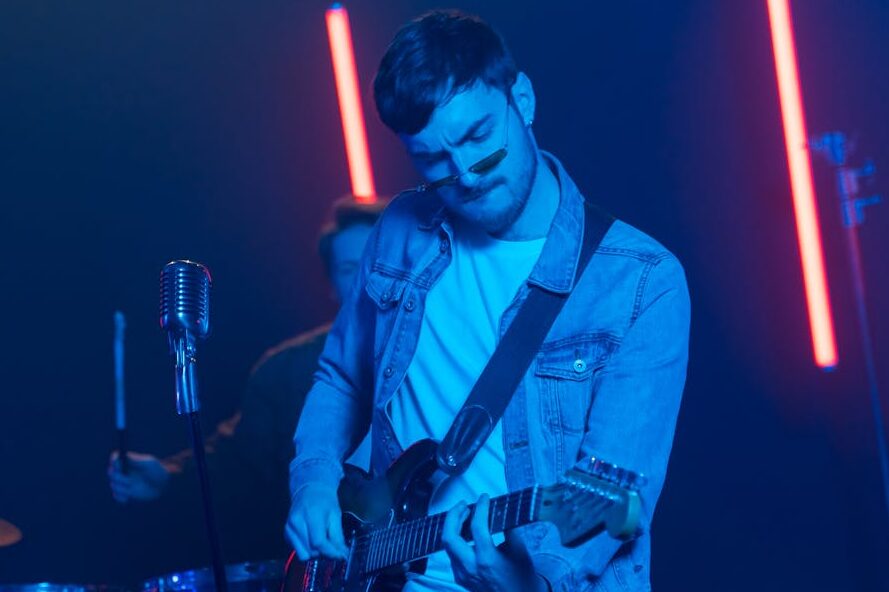 A musician passionately plays an electric guitar on stage with a band under vibrant lighting.