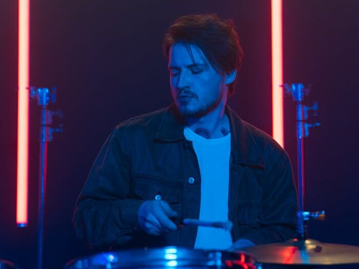 A male musician in a denim jacket playing drums under vibrant neon lights.